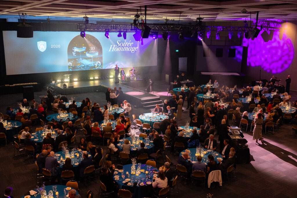 Vue aérienne de la salle 200 du Centre des congrès de Québec, aménagée avec des tables de banquets et une scène, avec des gens qui discutent lors de la soirée Hommage aux ambassadeurs 2026.